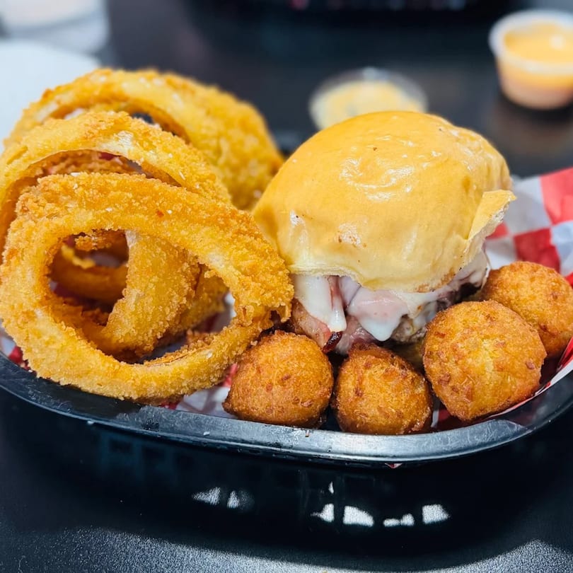 A Bergstrom's burger with melted cheese, onion rings, and hush puppies in a black basket.